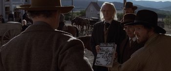 Movie still from “Unforgiven” (1992), directed by Clint Eastwood – A man in a suit and tie holding a comic book; Medium shot, Over the shoulder angle