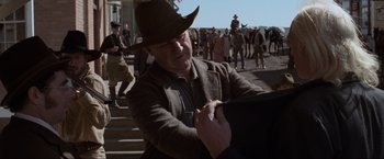 Movie still from “Unforgiven” (1992), directed by Clint Eastwood – A man wearing a hat is holding something up to his face; Medium shot, Over the shoulder angle