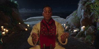 Movie still from “Vacation Friends” (2021), directed by Clay Tarver – A man sitting on the ground with a red scarf around his neck; Medium shot, Low angle