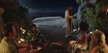 Movie still from “Vacation Friends” (2021), directed by Clay Tarver – A group of people sitting on the ground near the ocean; Wide shot, High angle