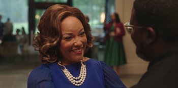 Movie still from “Vacation Friends” (2021), directed by Clay Tarver – An older woman wearing a blue dress and a pearl necklace; Close Up shot, Over the shoulder angle