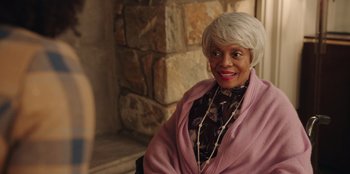 Movie still from “Vacation Friends” (2021), directed by Clay Tarver – An older woman with white hair sitting in front of a stone wall; Medium shot, Over the shoulder angle