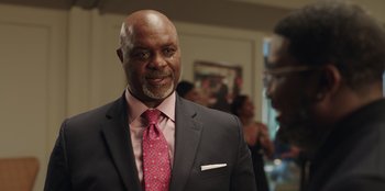 Movie still from “Vacation Friends” (2021), directed by Clay Tarver – A man in a suit and a pink tie smiling; Medium shot, Over the shoulder angle