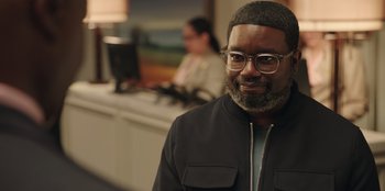 Movie still from “Vacation Friends” (2021), directed by Clay Tarver – A man with a beard wearing glasses and a black jacket; Medium shot, Over the shoulder angle