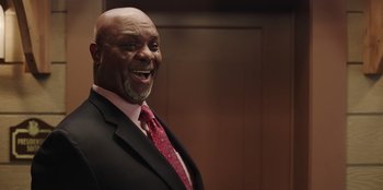 Movie still from “Vacation Friends” (2021), directed by Clay Tarver – A man in a suit and tie smiling for the camera; Medium shot, Low angle