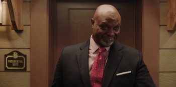 Movie still from “Vacation Friends” (2021), directed by Clay Tarver – A man in a suit and tie smiling for the camera; Medium shot, Low angle