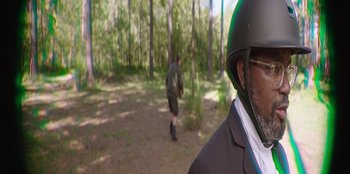 Movie still from “Vacation Friends” (2021), directed by Clay Tarver – A man in a suit and helmet in a wooded area; Medium shot, Over the shoulder angle