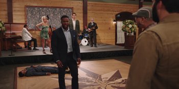 Movie still from “Vacation Friends” (2021), directed by Clay Tarver – A man standing on a stage in front of a group of people; Wide shot, Over the shoulder angle