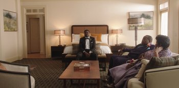 Movie still from “Vacation Friends” (2021), directed by Clay Tarver – A man sitting on a couch in front of a bed in a hotel room; Wide shot, High angle