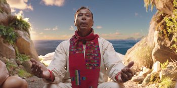 Movie still from “Vacation Friends” (2021), directed by Clay Tarver – A man sitting on the beach wearing a red and white scarf; Medium shot, Low angle