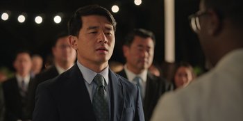 Movie still from “Vacation Friends 2” (2023), directed by Clay Tarver – A man in a suit and tie standing in front of other men; Close Up shot, Over the shoulder angle