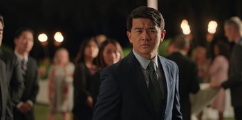 Movie still from “Vacation Friends 2” (2023), directed by Clay Tarver – A man wearing a suit and tie standing in front of a group of people; Medium shot, Over the shoulder angle