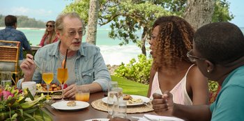 Movie still from “Vacation Friends 2” (2023), directed by Clay Tarver – A man and a woman sitting at a table with plates of food on it; Medium shot, Over the shoulder angle