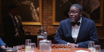 Movie still from “Vacation Friends 2” (2023), directed by Clay Tarver – A man sitting at a table in front of a row of whiskey glasses; Medium shot, Over the shoulder angle