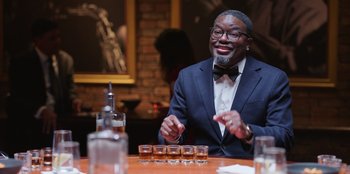 Movie still from “Vacation Friends 2” (2023), directed by Clay Tarver – A man sitting at a table in front of a row of glasses; Medium shot, Over the shoulder angle
