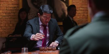 Movie still from “Vacation Friends 2” (2023), directed by Clay Tarver – A man sitting at a table holding a glass of whiskey; Medium shot, Over the shoulder angle