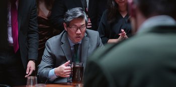 Movie still from “Vacation Friends 2” (2023), directed by Clay Tarver – A man in a suit and tie sitting at a table with a drink; Medium shot, Over the shoulder angle