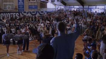 Movie still from “Varsity Blues” (1999), directed by Brian Robbins – A crowd of people in a gymnasium with a man holding his hand up; Wide shot, High angle