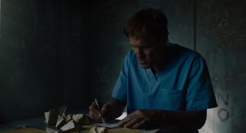 Movie still from “Venom: Let There Be Carnage” (2021), directed by Andy Serkis – A man writing on a piece of paper in a dark room; Medium shot, Low angle