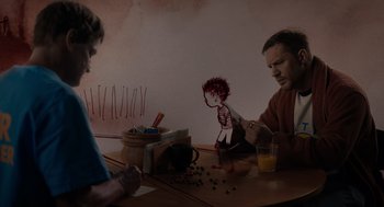 Movie still from “Venom: Let There Be Carnage” (2021), directed by Andy Serkis – Two people sitting at a wooden table with a paper doll; Medium shot, Over the shoulder angle