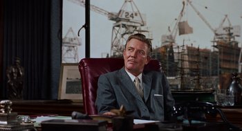 Movie still from “Vertigo” (1958), directed by Alfred Hitchcock – A man in a suit sitting at a desk in an office; Medium shot, Over the shoulder angle