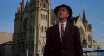 Movie still from “Vertigo” (1958), directed by Alfred Hitchcock – A man in a suit and hat standing in front of a church; Medium shot, Low angle