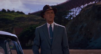 Movie still from “Vertigo” (1958), directed by Alfred Hitchcock – A man in a suit and hat standing in front of a bridge; Medium shot, Low angle