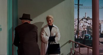 Movie still from “Vertigo” (1958), directed by Alfred Hitchcock – A woman in a white coat holding a black purse; Medium shot, Over the shoulder angle