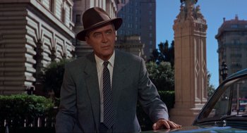 Movie still from “Vertigo” (1958), directed by Alfred Hitchcock – A man in a suit and hat standing in front of a building; Medium shot, Over the shoulder angle