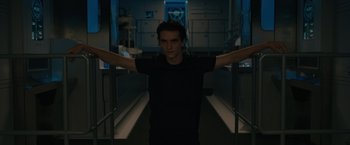 Movie still from “Voyagers” (2021), directed by Neil Burger – A young man standing in a dark room with his arms outstretched; Medium shot, Low angle