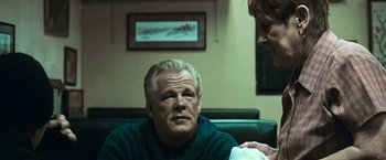 Movie still from “Warrior” (2011), directed by Gavin O'Connor – An older man sitting on a couch next to a woman; Medium shot, Over the shoulder angle