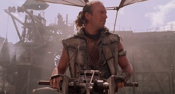 Movie still from “Waterworld” (1995), directed by Kevin Reynolds – A man in a leather vest is sitting on top of a boat; Medium shot, Low angle