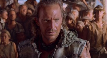 Movie still from “Waterworld” (1995), directed by Kevin Reynolds – A person with a bunch of people in the background; Close Up shot, Over the shoulder angle