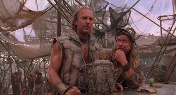 Movie still from “Waterworld” (1995), directed by Kevin Reynolds – Two men are dressed in barbarian garb and one is holding a drum; Medium shot, Over the shoulder angle