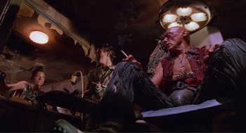 Movie still from “Waterworld” (1995), directed by Kevin Reynolds – A man smoking a cigarette next to another man sitting down; Medium shot, Low angle