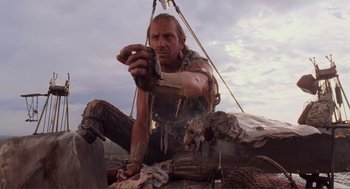 Movie still from “Waterworld” (1995), directed by Kevin Reynolds – A man sitting on top of a boat in the water; Medium shot, Low angle