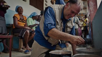 Movie still from “We Feed People” (2022), directed by Ron Howard – A group of people are gathered around a man cooking food; Medium shot, Over the shoulder angle