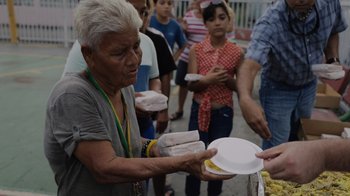 Movie still from “We Feed People” (2022), directed by Ron Howard – A group of people are holding plates of food; Medium shot, Over the shoulder angle