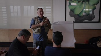 Movie still from “We Feed People” (2022), directed by Ron Howard – A group of people sitting in front of a man holding a cell phone; Medium shot, Over the shoulder angle