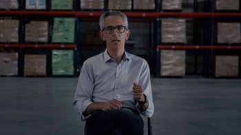 Movie still from “We Feed People” (2022), directed by Ron Howard – An older man in glasses and a white dress shirt is sitting in a warehouse; Medium shot, Over the shoulder angle
