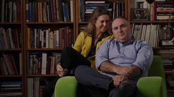 Movie still from “We Feed People” (2022), directed by Ron Howard – A man and a woman sitting on a couch in front of bookshelves; Medium shot, Low angle