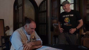 Movie still from “We Feed People” (2022), directed by Ron Howard – A man sitting at a table with another man standing next to him; Medium shot, Over the shoulder angle