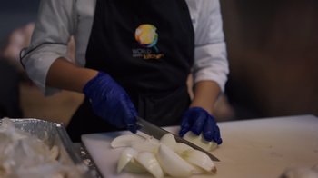 Movie still from “We Feed People” (2022), directed by Ron Howard – A person wearing blue gloves cutting onions on a cutting board; Extreme Close Up shot, Low angle