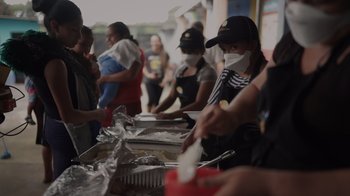 Movie still from “We Feed People” (2022), directed by Ron Howard – A group of people standing around a table with food on it; Medium shot, Over the shoulder angle