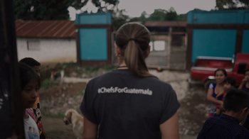 Movie still from “We Feed People” (2022), directed by Ron Howard – A woman wearing a shirt that says " chefs for guatemala " on the back of her shirt; Close Up shot, Over the shoulder angle