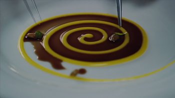 Movie still from “We Feed People” (2022), directed by Ron Howard – A swirl of melted chocolate and yellow sauce on top of a white plate; Extreme Close Up shot, Overhead angle