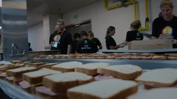Movie still from “We Feed People” (2022), directed by Ron Howard – A group of people standing in front of sandwiches on a table; Extreme Close Up shot, Low angle