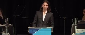 Movie still from “WeWork: Or the Making and Breaking of a $47 Billion Unicorn” (2021), directed by Jed Rothstein – A man standing at a podium in front of microphones; Medium shot, Low angle