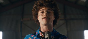 Movie still from “Weird: The Al Yankovic Story” (2022), directed by Eric Appel – A man with curly brown hair wearing glasses and holding a microphone; Close Up shot, Low angle