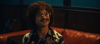 Movie still from “Weird: The Al Yankovic Story” (2022), directed by Eric Appel – A man sitting on top of a red couch wearing glasses; Close Up shot, Over the shoulder angle